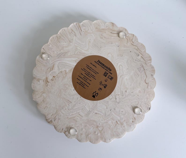 May include: A round, beige Jesmonite tray with a scalloped edge and a marbled pattern. The tray has a brown circular sticker with instructions and care information. Clear, round bumpers are attached to the bottom.