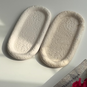 Może przedstawiać: Dwie owalne, kremowe ceramiczne tace o teksturowanej powierzchni. Tace leżą obok siebie na białej powierzchni. Tekst "HandMadebyAnyA" jest widoczny na szarej książce w prawym dolnym rogu.