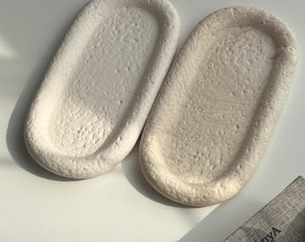 Puede incluir: Dos bandejas ovaladas de cerámica blanquecina con superficies texturizadas. Las bandejas están una al lado de la otra sobre una superficie blanca. El texto "HandMadebyAnyA" es visible en un libro gris en la esquina inferior derecha.