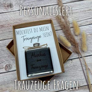 Może przedstawiać: Czarny skórzany flakon z srebrnymi krawędziami, spersonalizowany imieniem i słowem "Trauzeuge", znajduje się w kartonowym pudełku. Pudełko zawiera kartkę z niemieckim tekstem. Obraz zawiera suszone rośliny.