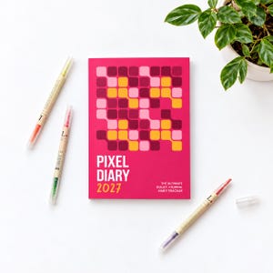 May include: A rectangular 2027 Pixel Diary bullet journal habit tracker with a bright pink cover featuring a grid of rounded squares in shades of pink, yellow, and dark red. Three dual-tip markers with clear caps are positioned around the journal.