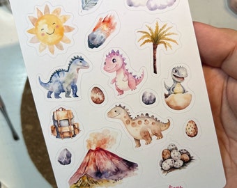 Op de afbeelding: Een vel met kleurrijke dinosaurus stickers met een gele zon, een roze dinosaurus, een blauwe dinosaurus, een bruine dinosaurus, een groene palmboom, een vulkaan, een bruine rugzak, een grijze wolk, een rode meteoor, een bruin ei en witte eieren. Het vel zegt "Fun Dino Stickers" en "Ink Hearted Creations".