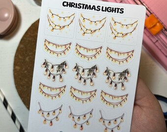 Puede incluir: Una hoja de pegatinas con varios diseños de luces de Navidad. Las pegatinas son blancas con un acabado brillante y presentan diferentes estilos de luces de cadena, incluyendo algunas con ramas de pino.