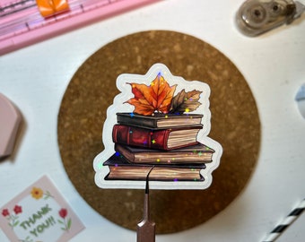 May include: A stack of four books with an autumn-coloured leaf on top. The books are decorated with glitter and have a holographic finish.