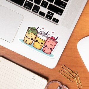 May include: A sticker depicting three cartoon bubble tea drinks with smiling faces. The drinks are in clear cups with various toppings: green tea, whipped cream, and strawberries. The sticker is on a laptop.