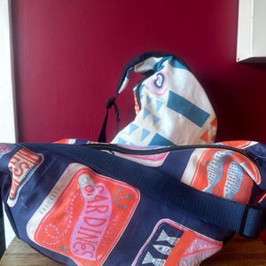 May include: A navy blue fanny pack and backpack with a sardine can pattern. The fanny pack has a navy blue strap and zipper. The backpack is white with blue, pink, and orange geometric shapes. The fanny pack has the word "Sardines" on it.