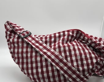 May include: A red and white checkered crossbody bag with an adjustable strap and a black buckle. The bag is made of a soft, woven fabric and has a zipper closure. The bag is designed to be worn across the body.