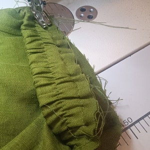May include: Close-up of a green fabric being sewn on a sewing machine. The fabric is gathered and pleated, creating a ruffled edge.