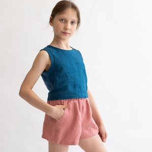 May include: A young person wearing a blue sleeveless top and pink linen shorts. The top has a round neckline and the shorts have an elastic waistband.