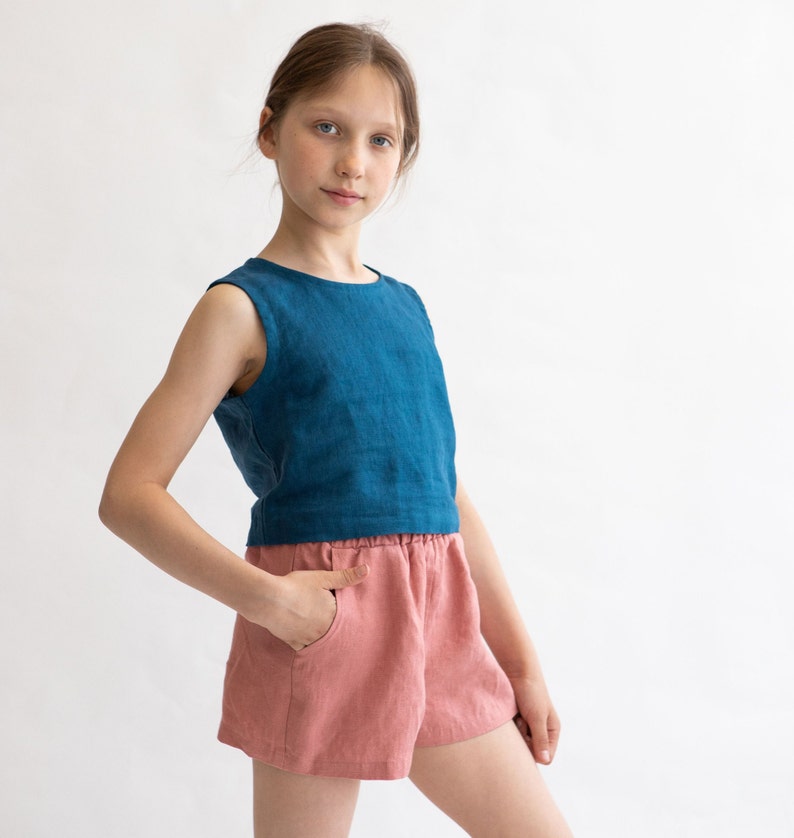 May include: A young person wearing a blue sleeveless top and pink linen shorts. The top has a round neckline and the shorts have an elastic waistband.