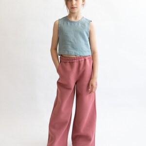 May include: A young person wearing a light blue sleeveless top and wide-legged, dusty rose pants.