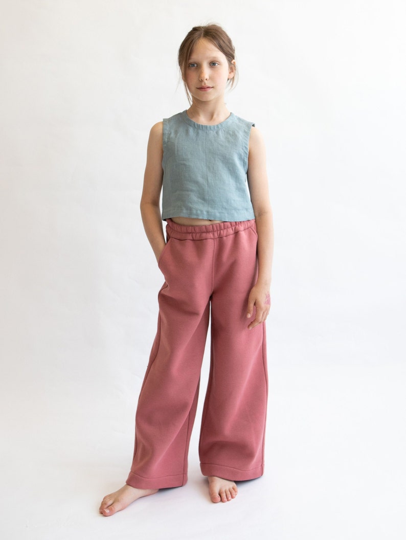 May include: A young person wearing a light blue sleeveless top and wide-legged, dusty rose pants.