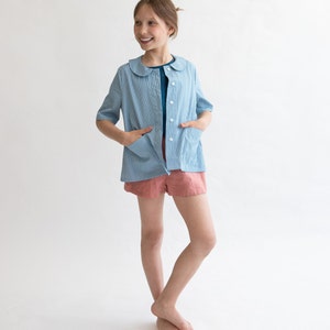 May include: A blue and white striped shirt with a peter pan collar and short sleeves. The shirt has a button-up front and two pockets. The model is wearing the shirt with a teal blue undershirt and pink shorts.