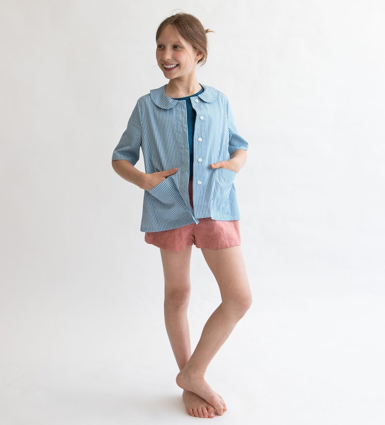 May include: A blue and white striped shirt with a peter pan collar and short sleeves. The shirt has a button-up front and two pockets. The model is wearing the shirt with a teal blue undershirt and pink shorts.