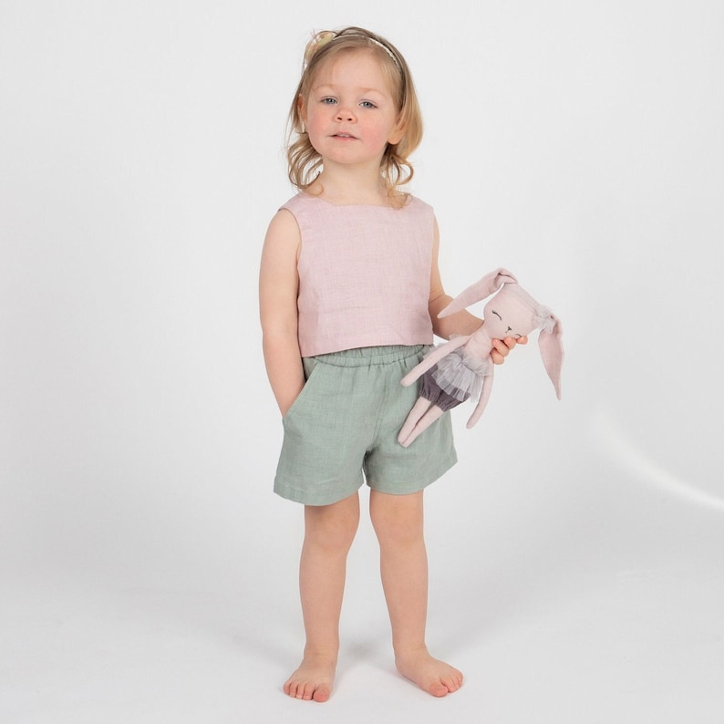 May include: A young girl wearing a pink linen tank top and green linen shorts holds a pink fabric bunny doll.