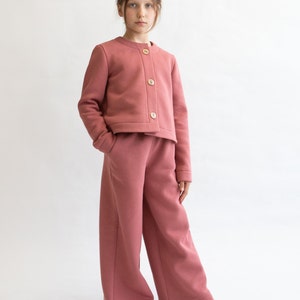 May include: A young person wearing a pink cropped cardigan with button closures and matching wide-leg pants.