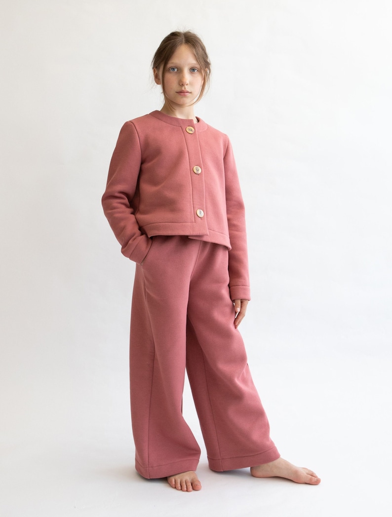 May include: A young person wearing a pink cropped cardigan with button closures and matching wide-leg pants.
