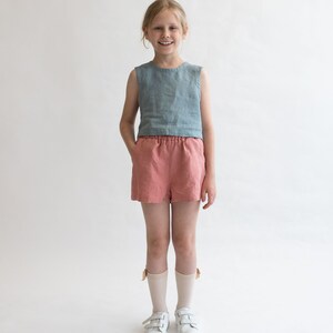 May include: A young person wearing a light blue sleeveless linen top and coral linen shorts. They are wearing white sneakers and white socks with a light brown bow detail.