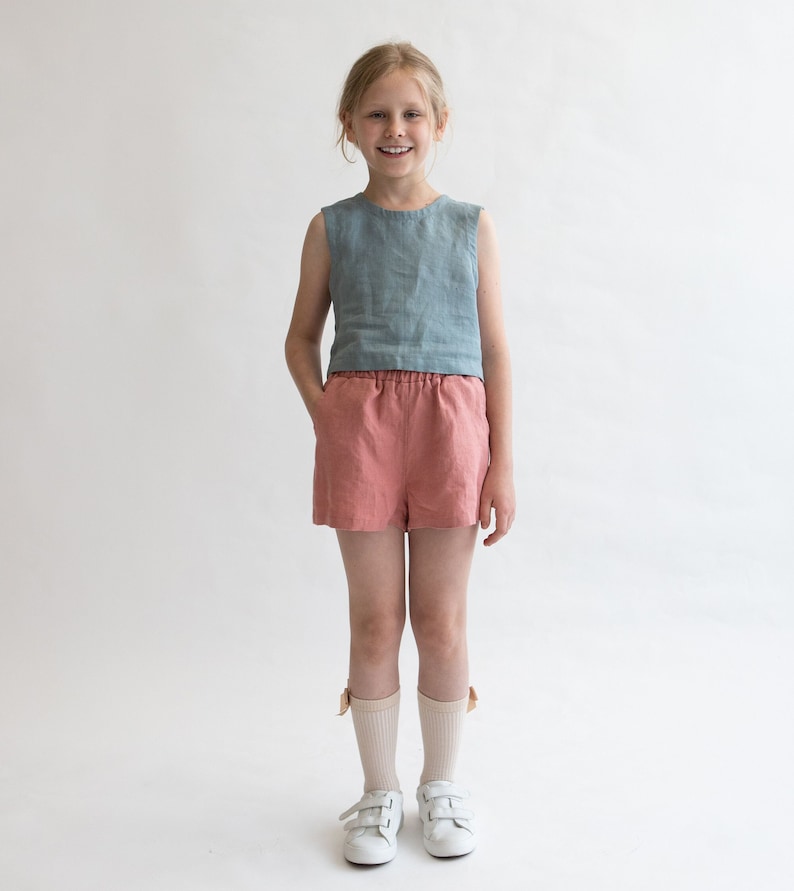 May include: A young person wearing a light blue sleeveless linen top and coral linen shorts. They are wearing white sneakers and white socks with a light brown bow detail.