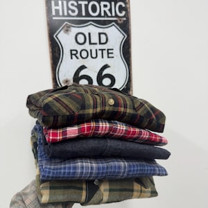Könnte beinhalten: Ein Stapel karierter Flanellhemden in verschiedenen Farben, darunter Grün, Rot, Blau und Grau. Die Hemden sind gefaltet und übereinander gestapelt. Im Hintergrund befindet sich ein Vintage-Route 66-Schild.