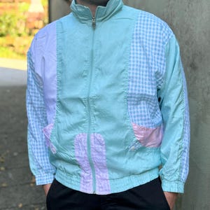 Op de afbeelding: Een lichtblauw jack met een wit en blauw ruitpatroon aan de ene kant en een roze en witte streep aan de andere kant.