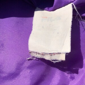 May include: A close-up of a purple fabric with a white tag that says "Fila" in a faded pink font.
