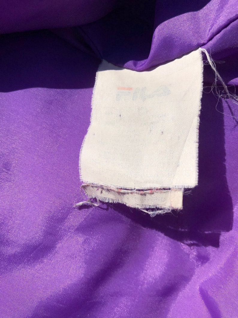 May include: A close-up of a purple fabric with a white tag that says "Fila" in a faded pink font.