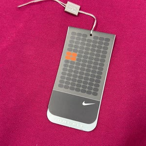 May include: A magenta-colored garment with a tag attached. The tag is gray with a grid pattern, orange squares, and the Nike logo. The tag reads "DRI-FIT". A white string and a small silver square are attached to the tag.