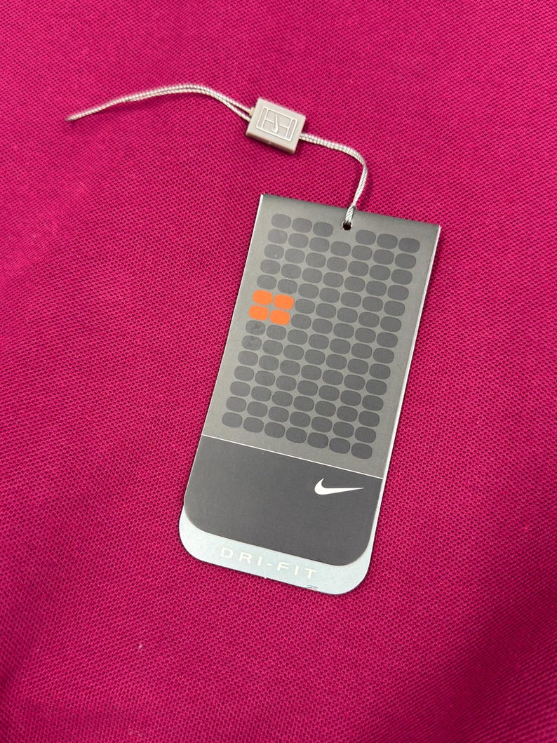 May include: A magenta-colored garment with a tag attached. The tag is gray with a grid pattern, orange squares, and the Nike logo. The tag reads "DRI-FIT". A white string and a small silver square are attached to the tag.