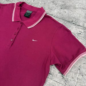 May include: A magenta polo shirt with a white and pink striped collar and sleeve trim. The shirt has a three-button placket and a small white Nike logo on the chest.