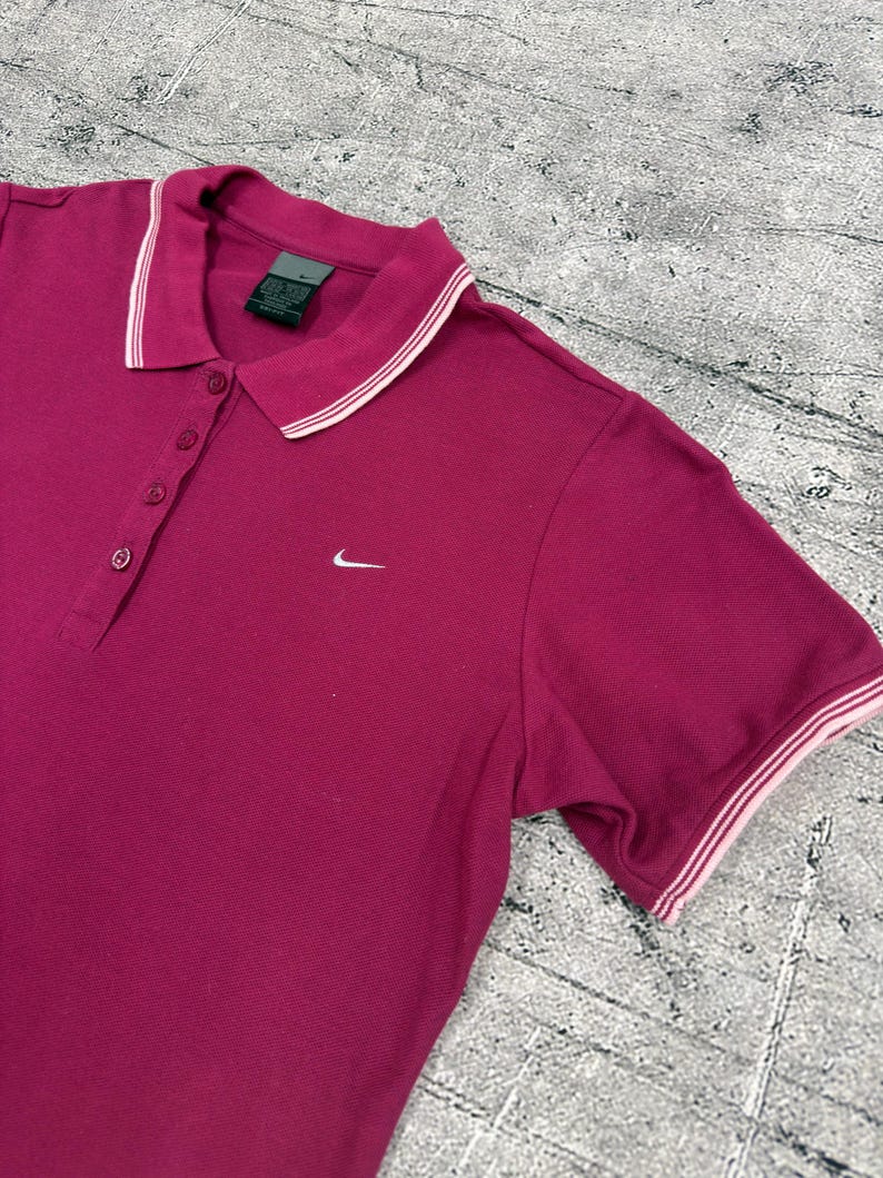 May include: A magenta polo shirt with a white and pink striped collar and sleeve trim. The shirt has a three-button placket and a small white Nike logo on the chest.