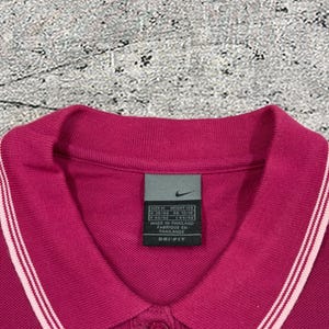 May include: A magenta Nike polo shirt with a collared neckline and a three-button placket. The collar and sleeve cuffs have white stripes. A Nike tag is visible near the collar. The shirt is made of Dri-FIT material.