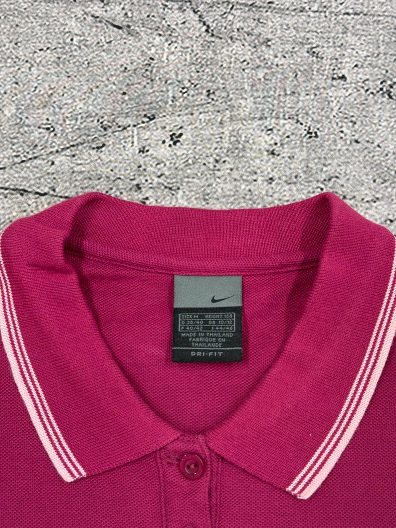 May include: A magenta Nike polo shirt with a collared neckline and a three-button placket. The collar and sleeve cuffs have white stripes. A Nike tag is visible near the collar. The shirt is made of Dri-FIT material.