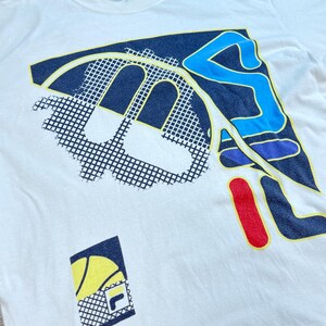 May include: Off-white t-shirt with a large graphic design. The design features a combination of navy blue, yellow, red, and light blue shapes, including a stylized letter "F" and a tennis ball. The shirt has a crew neck.