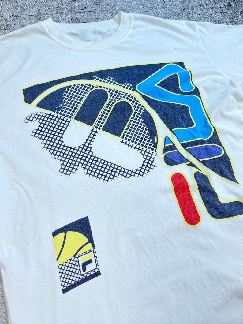 May include: Off-white t-shirt with a large graphic design. The design features a combination of navy blue, yellow, red, and light blue shapes, including a stylized letter "F" and a tennis ball. The shirt has a crew neck.