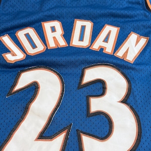 K&ouml;nnte beinhalten: Blaues Basketballtrikot mit dem Namen "JORDAN" in wei&szlig;en Buchstaben mit orangefarbenen Umrissen. Die Nummer "23" wird in gro&szlig;en wei&szlig;en Ziffern mit schwarzem und orangefarbenem Besatz angezeigt. Das Trikot hat einen blauen und orangefarbenen Ausschnitt.