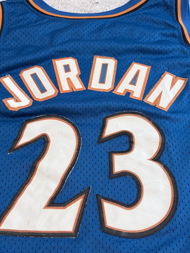 K&ouml;nnte beinhalten: Blaues Basketballtrikot mit dem Namen "JORDAN" in wei&szlig;en Buchstaben mit orangefarbenen Umrissen. Die Nummer "23" wird in gro&szlig;en wei&szlig;en Ziffern mit schwarzem und orangefarbenem Besatz angezeigt. Das Trikot hat einen blauen und orangefarbenen Ausschnitt.