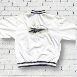 Op de afbeelding: Witte windbreaker-jas met een zwart-wit gestreepte kraag en manchetten. De jas heeft een grote, doorzichtige plastic patch op de rug met een zwart Reebok-logo.
