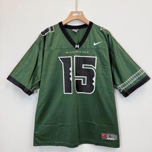 May include: Green football jersey with the number 15 in white, and the word "WARRIORS" below the neckline. The jersey has black trim on the sleeves and neckline, and a Nike logo. The jersey is hanging on a wooden hanger.