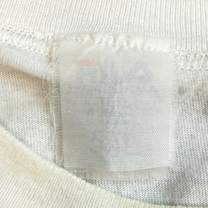 May include: Close-up of a white t-shirt with a rectangular tag sewn into the neckline. The tag is white and appears to have faded or blurred text. The t-shirt fabric has a ribbed texture.