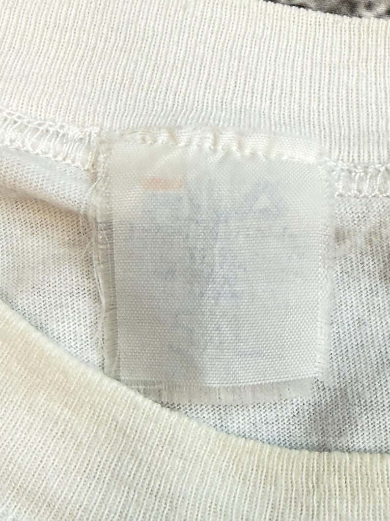 May include: Close-up of a white t-shirt with a rectangular tag sewn into the neckline. The tag is white and appears to have faded or blurred text. The t-shirt fabric has a ribbed texture.