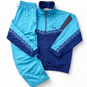 Könnte beinhalten: Ein zweiteiliger Fila-Trainingsanzug in Blautönen. Die Jacke hat einen Reißverschluss, einen Stehkragen und ein geometrisches Design auf den Ärmeln. Die Hose ist einfarbig hellblau mit Kordelzug. Das Fila-Logo ist auf beiden Teilen sichtbar.