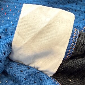 K&ouml;nnte beinhalten: Nahaufnahme eines blauen Mesh-Stoffs mit einem wei&szlig;en Etikett. Das Etikett enth&auml;lt Text. Der Stoff hat kleine L&ouml;cher und ist mit orangefarbenem Faden gen&auml;ht. Der Rand eines schwarzen Stoffes ist sichtbar.