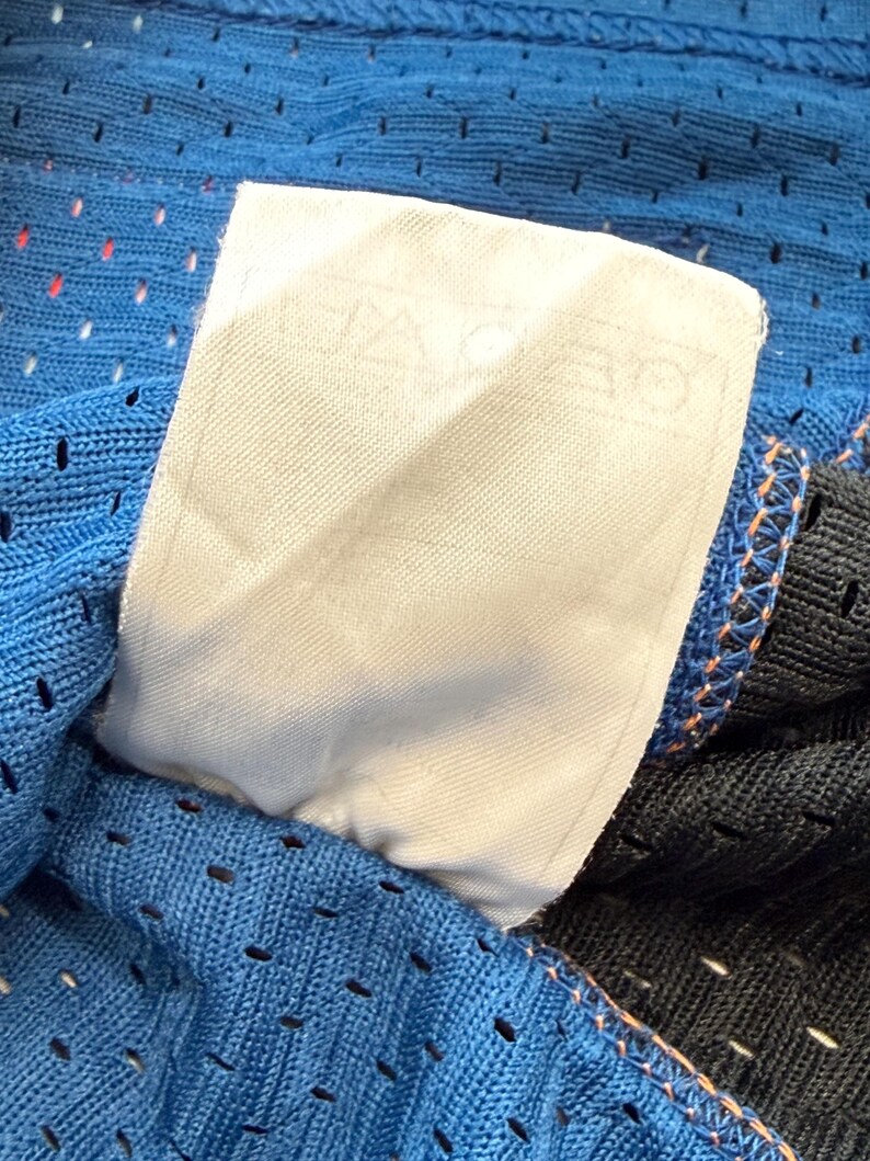 K&ouml;nnte beinhalten: Nahaufnahme eines blauen Mesh-Stoffs mit einem wei&szlig;en Etikett. Das Etikett enth&auml;lt Text. Der Stoff hat kleine L&ouml;cher und ist mit orangefarbenem Faden gen&auml;ht. Der Rand eines schwarzen Stoffes ist sichtbar.