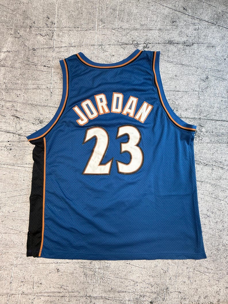 K&ouml;nnte beinhalten: Blaues Basketballtrikot mit dem Namen "JORDAN" und der Nummer "23" in Wei&szlig;. Das Trikot hat orangefarbene und schwarze Akzente. Das Trikot besteht aus einem Mesh-Material.