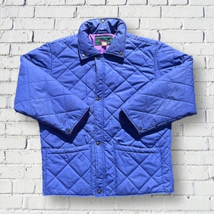 May include: A blue quilted jacket with a snap closure and two front pockets. The jacket has a purple lining and a green and pink label with the text "Trailblazer" on it.