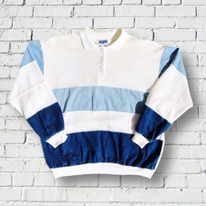 Può includere: Una camicia bianca a maniche lunghe con pannelli a righe blu e bianche e pannelli in denim blu. La camicia ha un colletto abbottonato e una chiusura con un solo bottone.
