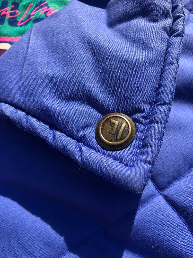 May include: Close-up of a blue quilted jacket with a gold metal button featuring the letter "T".