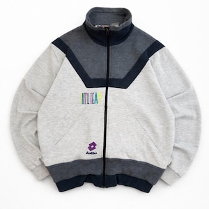 Puede incluir: Chaqueta deportiva gris con cremallera y cuello alto. La chaqueta presenta detalles en azul marino y gris oscuro, el texto "ITL TEA" en colores arcoíris y el logotipo de Lotto. La chaqueta está hecha de un material suave y cómodo.