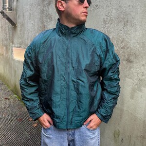 Puede incluir: Una chaqueta verde azulada con cierre de cremallera y cuello alto. La chaqueta está hecha de un material brillante e iridiscente.
